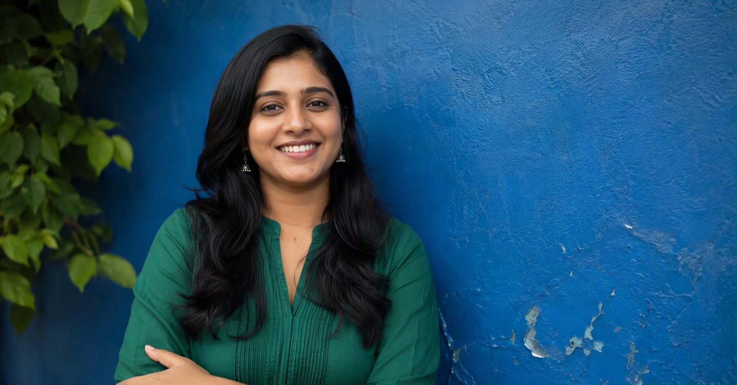 A smiling woman with brown skin wears an emerald green kurta and silver earrings in front of a deep blue wall, in soft shade.